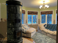 A Siberia green serpentine stone heater serving as the centerpiece of a sunlit living room with bay windows in a cabin in the woods.
